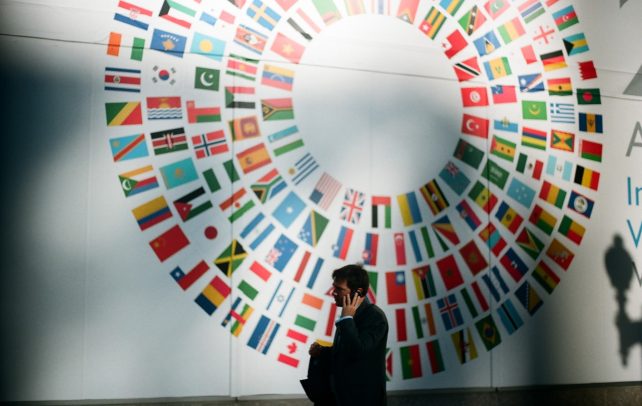 Homem de fato ao telemóvel em frente a parede com circulo com bandeiras do mundo inteiro