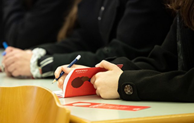 Sala de aula, imagem close up de braços em cima da  mesa, uma das pessoas com um lápis e a abrir caderninho vermelho com ilustração de mulher negra
